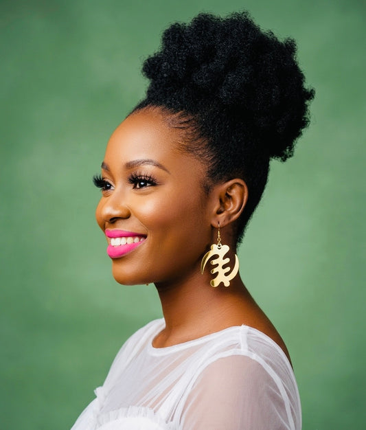 Woman wearing gold Gye Nyame dangling earrings against green background