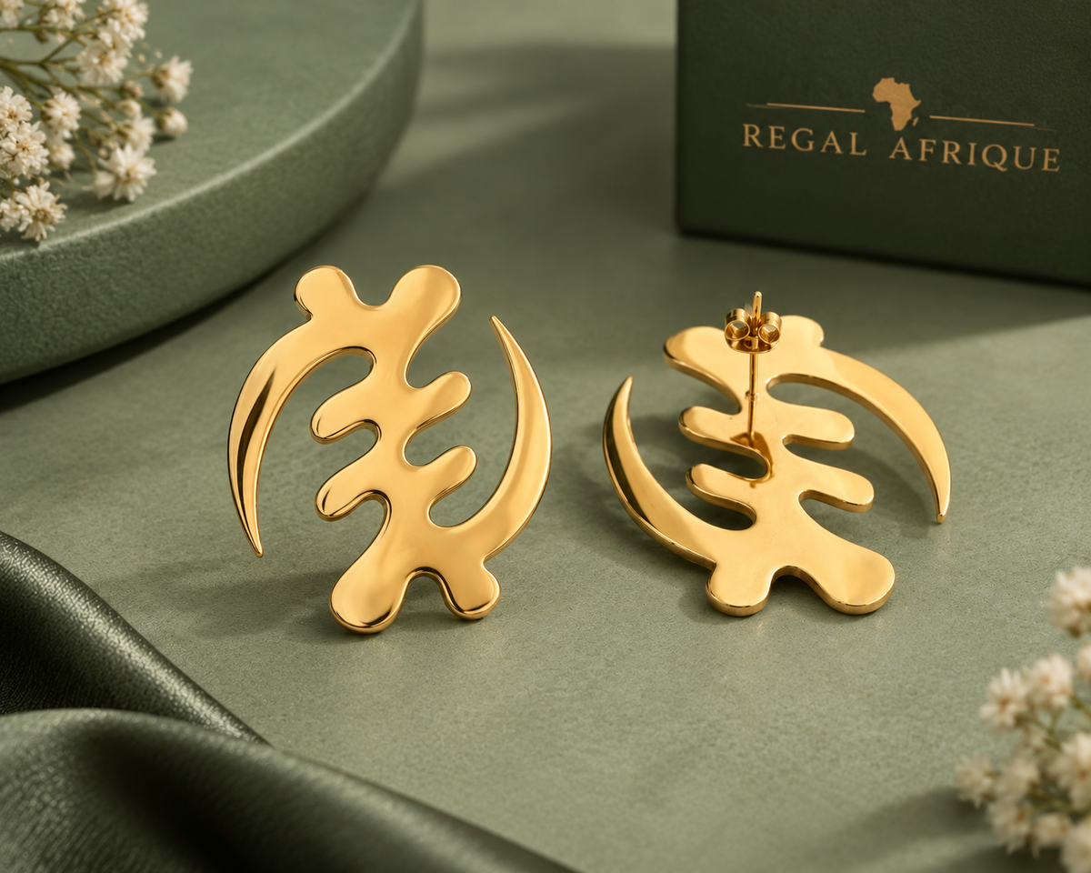 Large gold Gye Nyame stud earrings arranged on sage-green fabric beside a green box